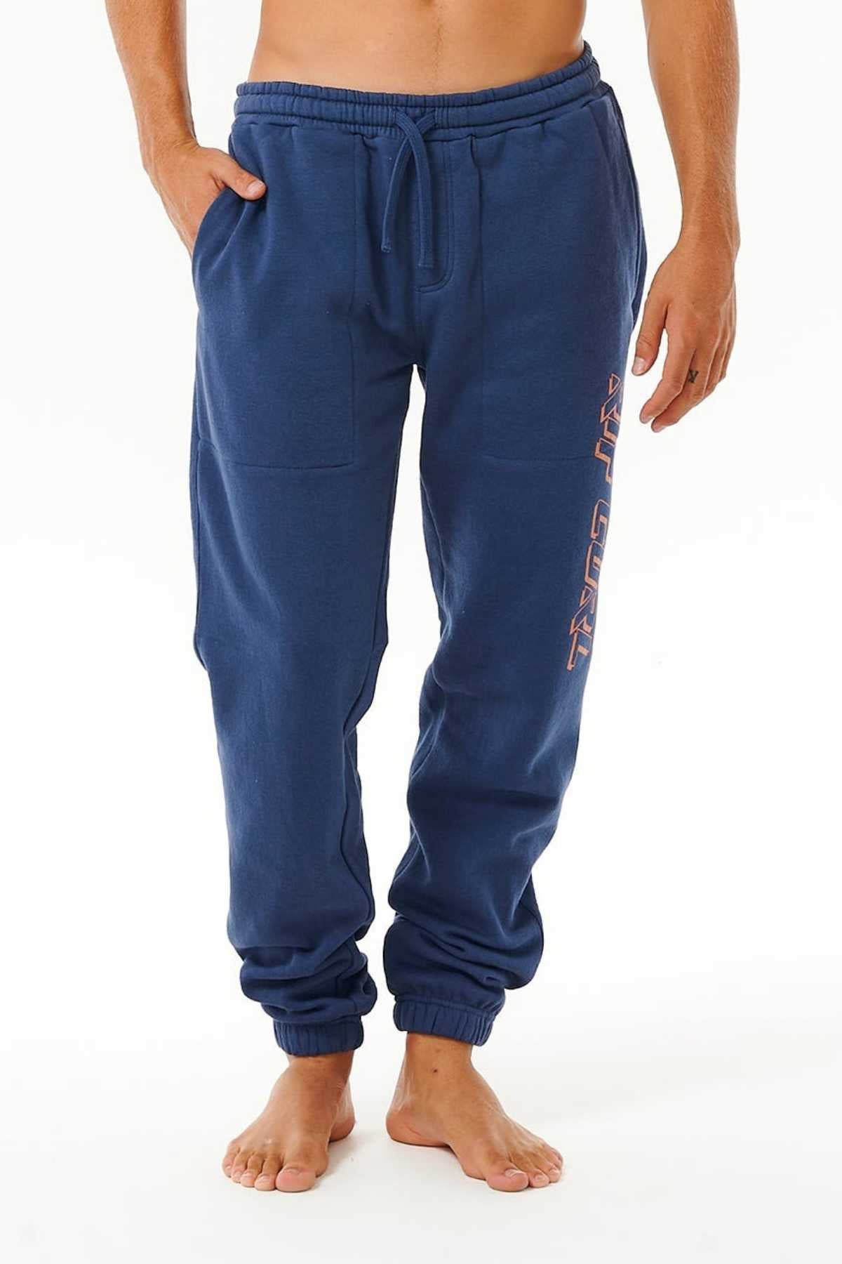 A person wears blue Rip Curl Icons Of Surf Trackpant joggers with pockets and a drawstring waist, text on the upper left leg, and stands barefoot against a white background.