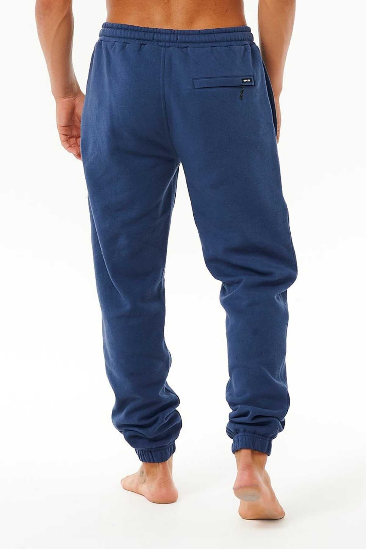 A barefoot man stands on a white background, waist down, wearing blue Rip Curl Icons Of Surf Trackpant sweatpants made from organic cotton with an elastic waistband, side pockets, and a small zippered back pocket.