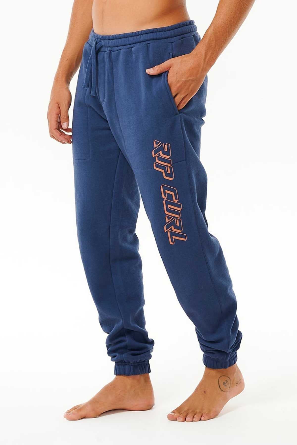 A person models blue Rip Curl Icons Of Surf Trackpant joggers featuring "RIP CURL" down one leg, made from organic cotton. The pants have a drawstring waist, elastic ankle cuffs, and are shown barefoot against a plain white background.