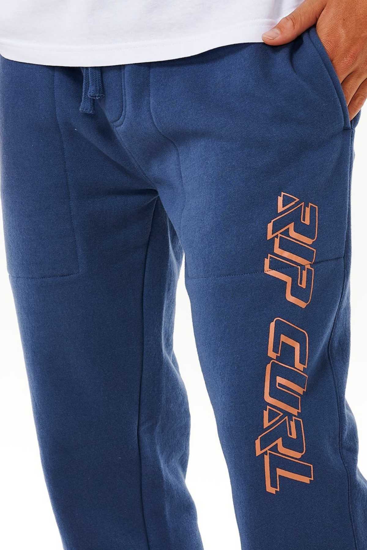 Wearing blue Rip Curl Icons Of Surf Trackpant sweatpants with bold orange "RIP CURL" down the left leg, made from organic cotton. Paired with a white shirt, one hand tucked in a pocket—a stylish and sustainable choice.