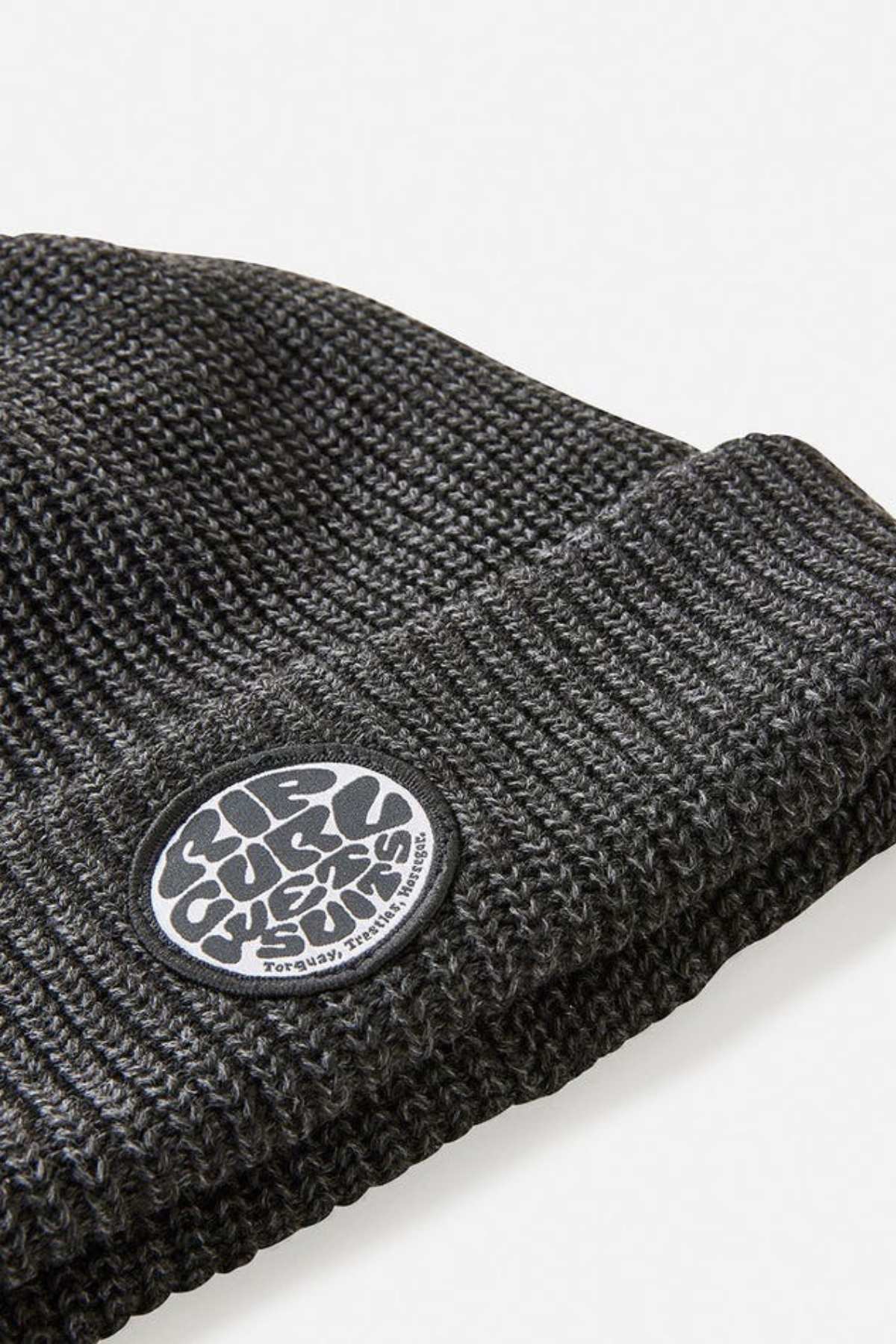 Close-up of the Rip Curl Icons Reg Beanie in dark gray, featuring a folded brim and a round black-and-white patch on the cuff. Made from recycled polyester, it's ideal for outdoor adventures.