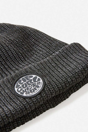 Close-up of the Rip Curl Icons Reg Beanie in dark gray, featuring a folded brim and a round black-and-white patch on the cuff. Made from recycled polyester, it's ideal for outdoor adventures.