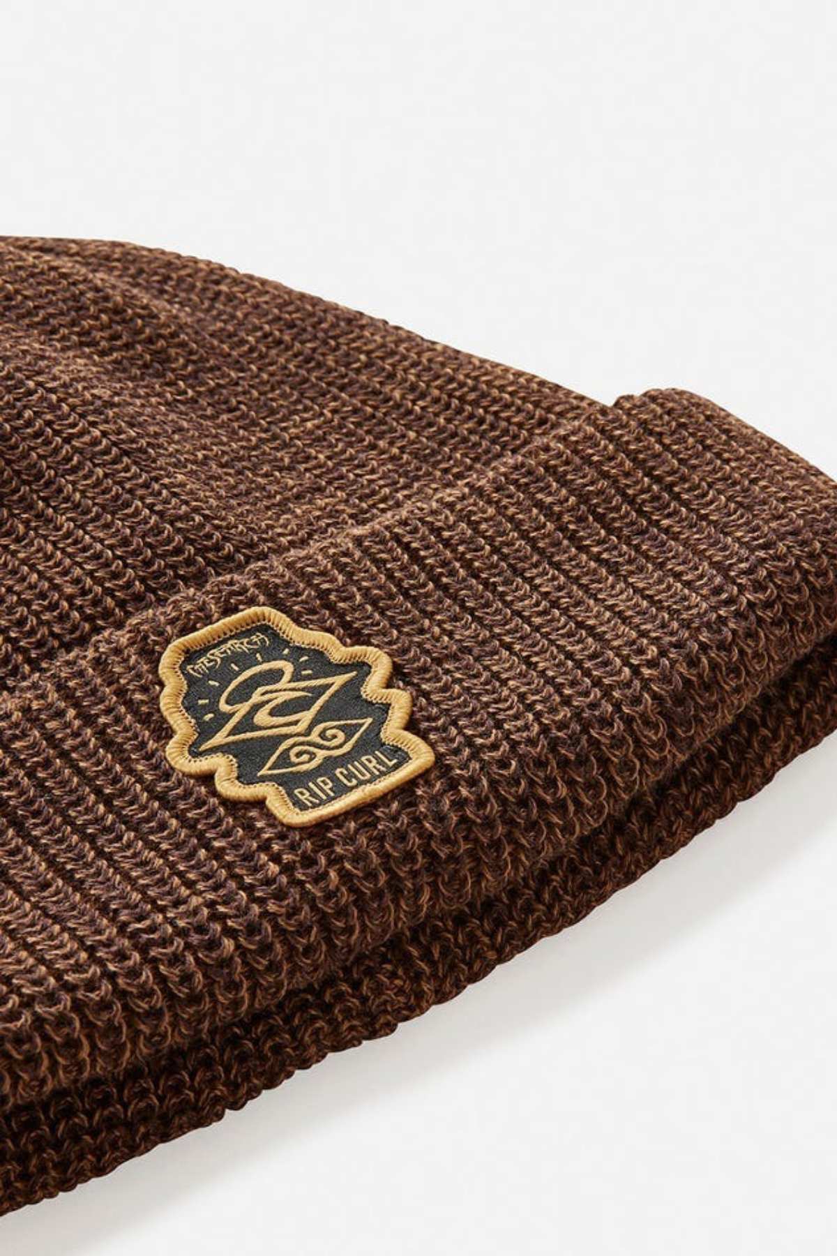 A close-up shot of the Rip Curl Icons Reg Beanie highlights its brown hue and intricate rectangular patch emblazoned with the "Rip Curl" logo. Made from recycled polyester, this outdoor adventure beanie features a fashionable folded brim and is set against a plain white backdrop.