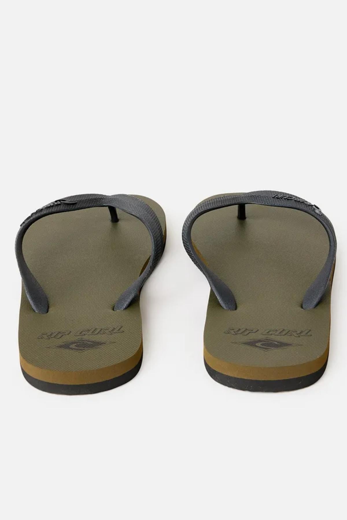 A pair of Rip Curl Icons of Surf Bloom Thongs in olive green with dark gray straps is displayed against a plain white background. These flip-flops feature a simple, casual design with the brand name "Rip Curl" imprinted on the footbeds near the heel area.