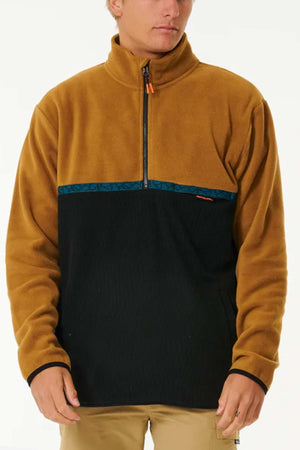 A person is wearing a Rip Curl Journey Polar Fleece Crew with a high collar and half zip front. The upper half is brown, the lower half black with a contrasting blue trim at the division, and it features a handy chest pocket. They are also wearing beige pants. The background is plain white.