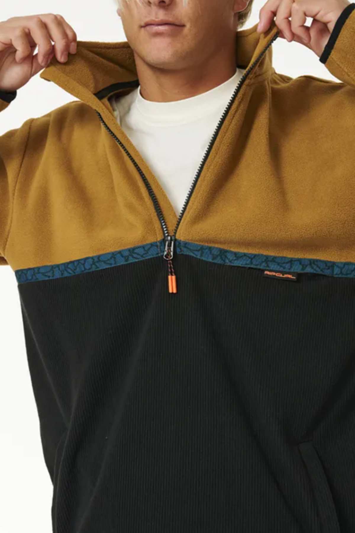 A person wearing a two-toned Rip Curl Journey Polar Fleece Crew with a mustard-yellow collar, upper chest, and sleeves, and a black lower section featuring a dark blue horizontal stripe near the half zip front. The person is partially zipping up the jacket, revealing a white undershirt beneath.