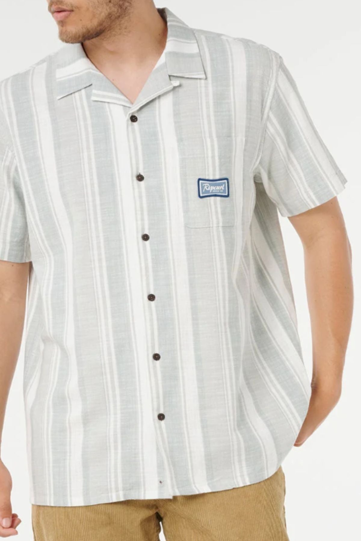 A man wearing the Rip Curl Mod Cali Stripe Short Sleeve Shirt with a small blue chest patch and tan corduroy pants stands with one hand in his pocket.