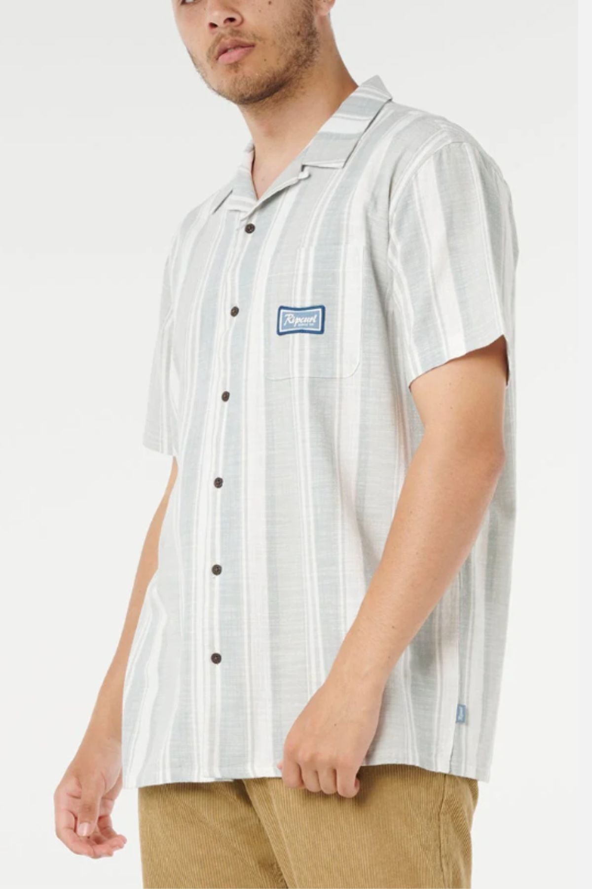 A man wears the Rip Curl Mod Cali Stripe Short Sleeve Shirt in light gray and white stripes with a small chest patch, paired with light brown pants, standing against a plain white background.