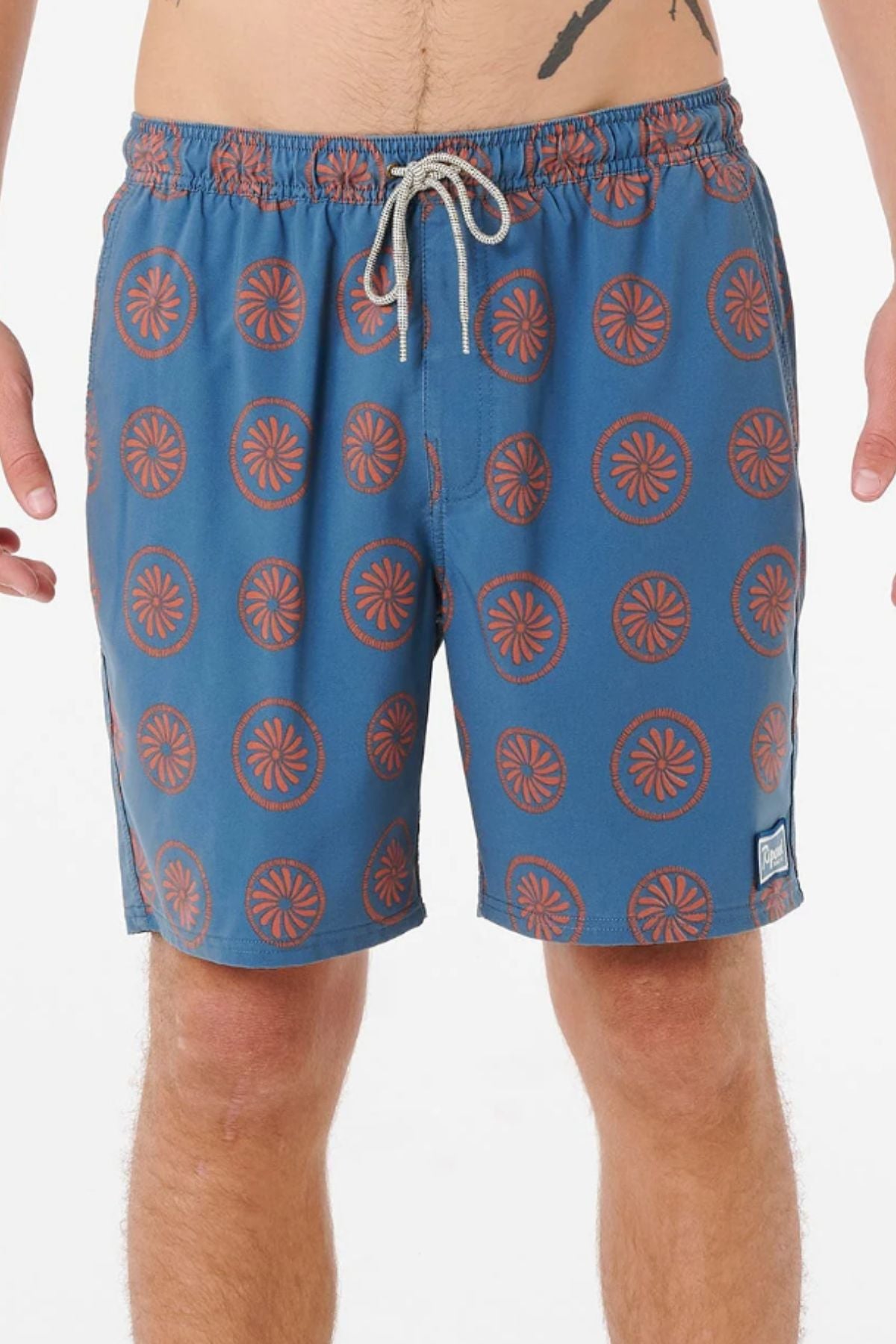 A person wears Rip Curl Mod Cali Volley Shorts in blue with an orange circular pattern, a white drawstring, and a small left-leg patch. Made from quick-dry fabric. The photo is cropped from mid-torso to above the knees.