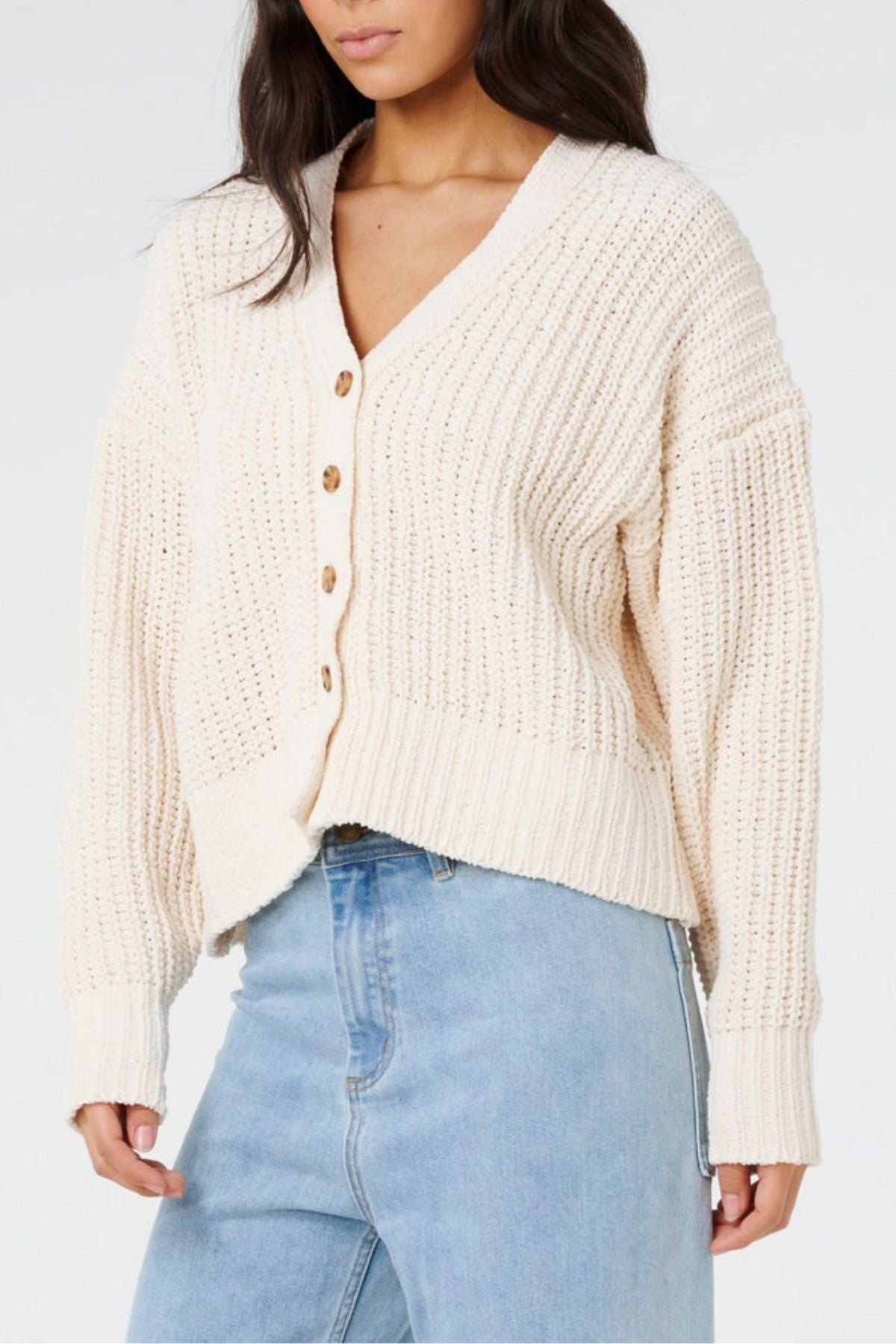 A woman wears the Rip Curl Neon Isles Cardigan—a cream textured knit with brown buttons—and light blue high-waisted jeans, standing against a plain background.