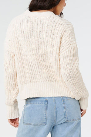 A person is seen from the back wearing the Rip Curl Neon Isles Cardigan in cream, styled with light blue high-waisted jeans. Their long dark hair contrasts against a plain white background.