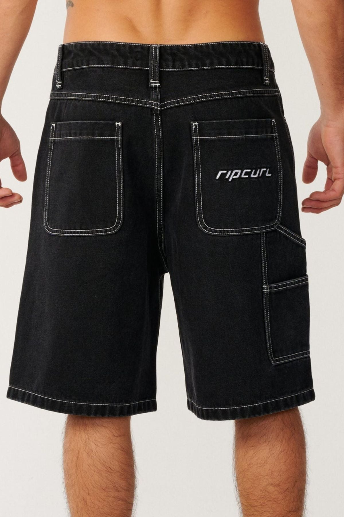 A person wearing black Rip Curl Nu Dose Denim Walkshorts with white stitching, viewed from the back. The shorts feature large back pockets and additional side pockets, giving them a relaxed skate style vibe.