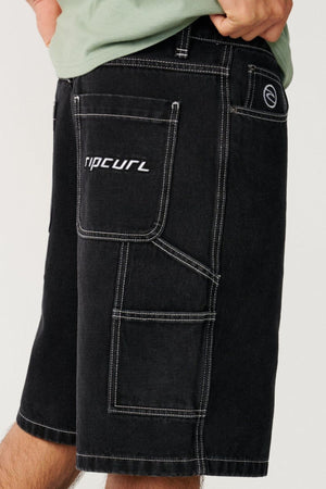 Wearing the Rip Curl Nu Dose Denim Walkshort, which features a black color with white stitching and a "ripcurl" logo on the back pocket, this person shows off its baggy skate-inspired silhouette with one hand in a side pocket. A glimpse of a green shirt is also visible.