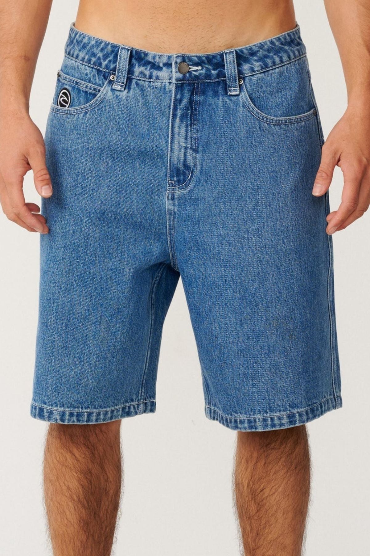 A person wearing the Rip Curl Nu Dose Denim Walkshort stands against a plain light background. The baggy fit shorts feature classic pockets, belt loops, and a visible button, with a small logo on the left pocket that channels subtle skate style. Only the torso and upper legs are visible.