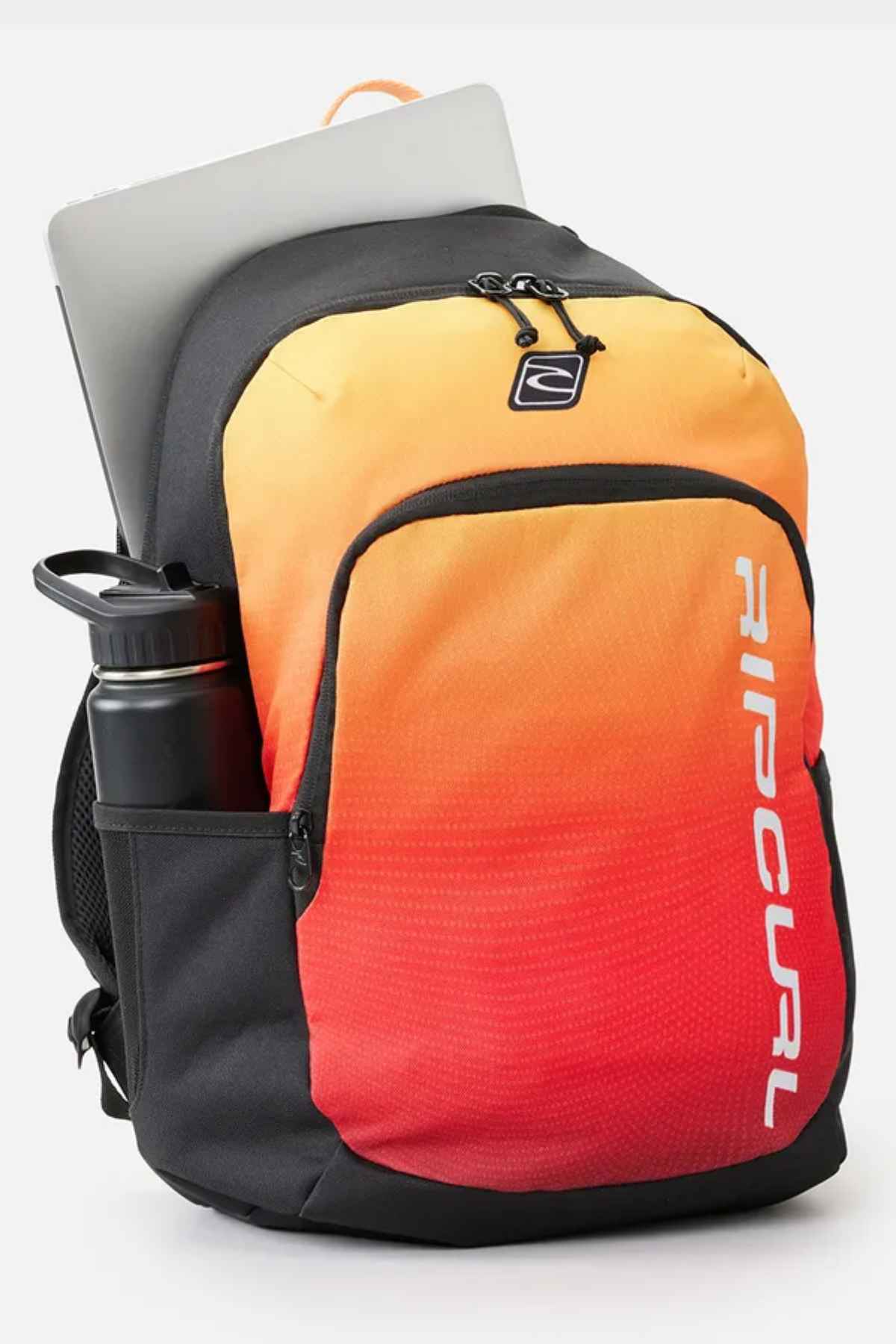 The Rip Curl Ozone 30L Back To School Backpack, with its gradient orange and red color scheme highlighted by black detailing, is ideal for back-to-school needs. It includes a spacious 30L capacity, featuring a water bottle conveniently placed in the side pocket and a laptop partially visible from the top. The front showcases the brand logo in a vertical orientation.