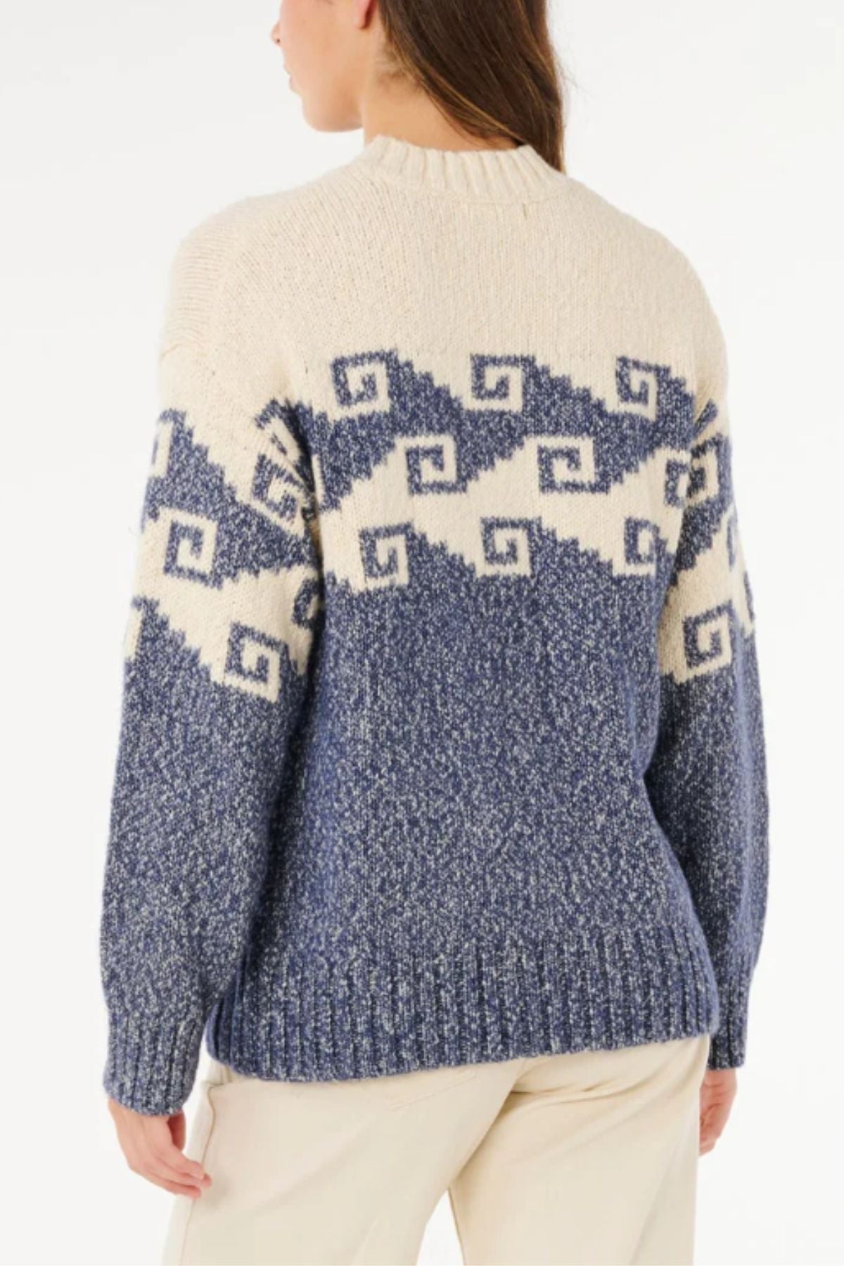 A person with long brown hair wears light-colored pants and the Rip Curl Pacific Coast Sweater in cream and blue, featuring a geometric zigzag pattern, standing with their back to the camera to display its coastal style.