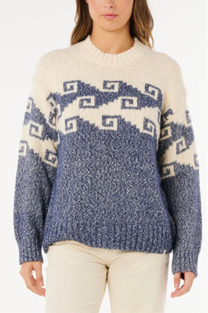 A person wears the Rip Curl Pacific Coast Sweater—cream and blue jacquard knit with a geometric square spiral pattern—paired with light-colored pants, standing against a plain white background.