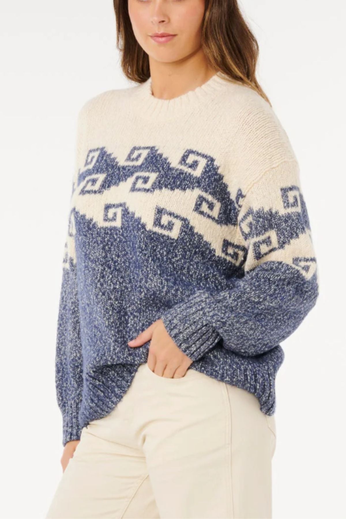 A woman wears the Rip Curl Pacific Coast Sweater, a cozy blue and cream jacquard knit with geometric patterns, paired with cream-colored pants. She stands against a white background with one hand in her pocket.