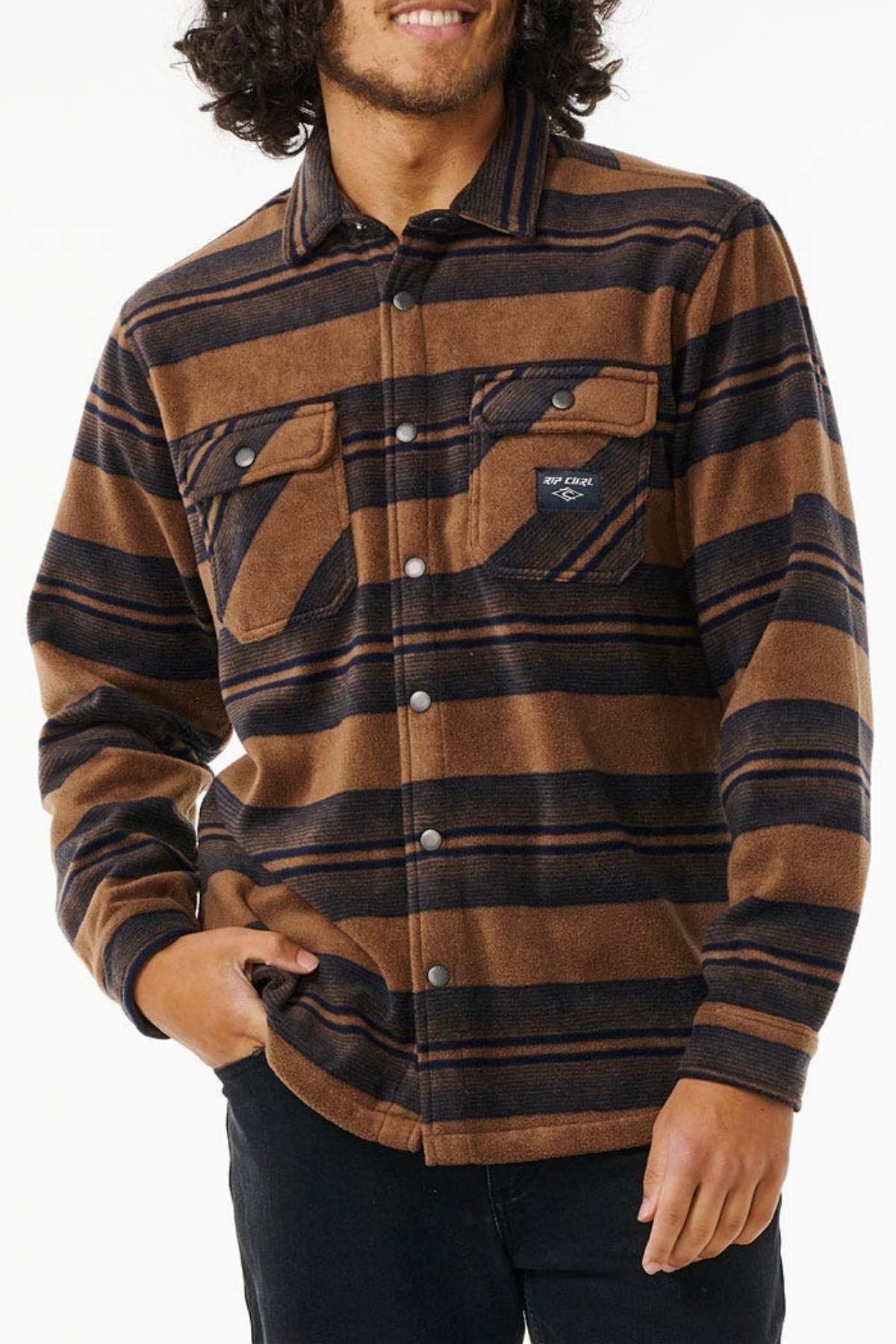 A person is wearing a brown and navy blue striped Rip Curl Party Pack Polar Fleece Shirt with chest pockets, standing with one hand in their pocket. The shirt has a casual, warm appearance, suitable for cooler weather. Made from recycled polyester, it's perfect for both style and sustainability. The person's face is partially visible, displaying a smile.