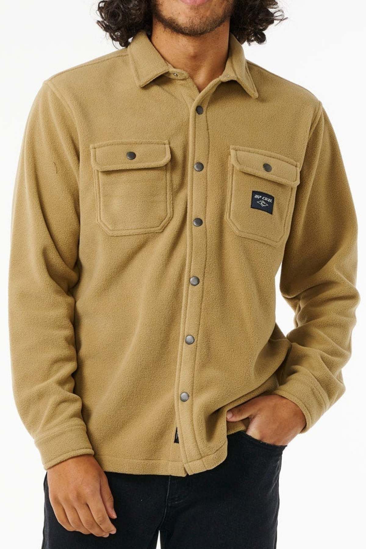 A person with curly hair is wearing a light brown, long-sleeve Rip Curl Party Pack Polar Fleece Shirt, featuring two chest pockets and a small black patch on one pocket. With their left hand in their pants pocket, they pose in front of a plain white background, making it an ideal look for cooler weather.