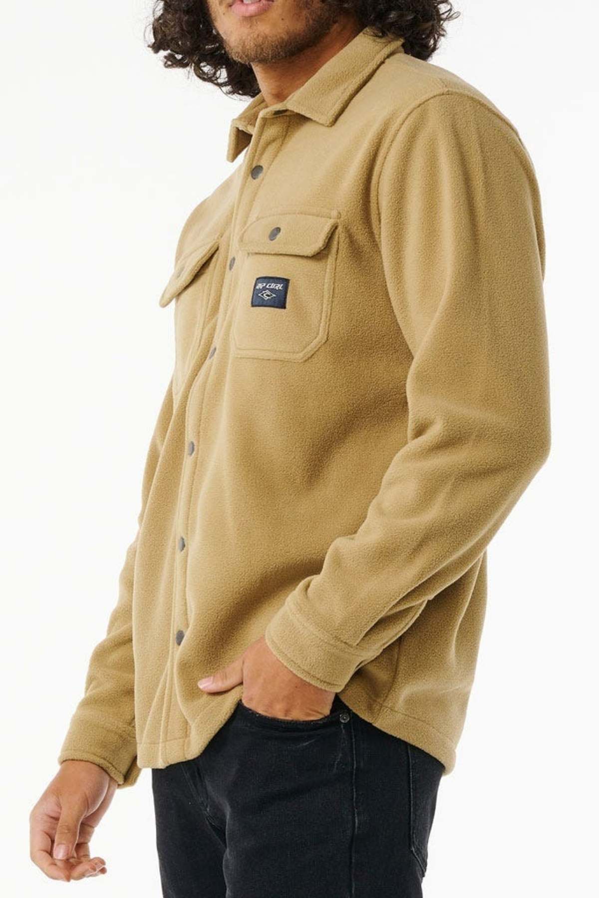 A person with shoulder-length hair is wearing a tan Rip Curl Party Pack Polar Fleece Shirt, featuring snap buttons and dual chest pockets, crafted from recycled polyester. The individual has one hand in the pocket of their dark-colored pants. The plain, light background suggests cooler weather.