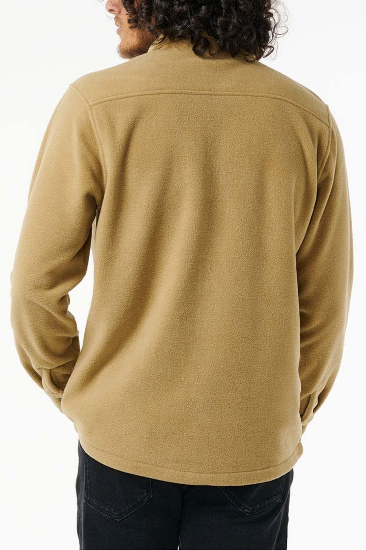 A person with long dark curly hair is seen from behind wearing a light brown Party Pack Polar Fleece Shirt by Rip Curl, made from recycled polyester, along with black jeans. The background is plain white.