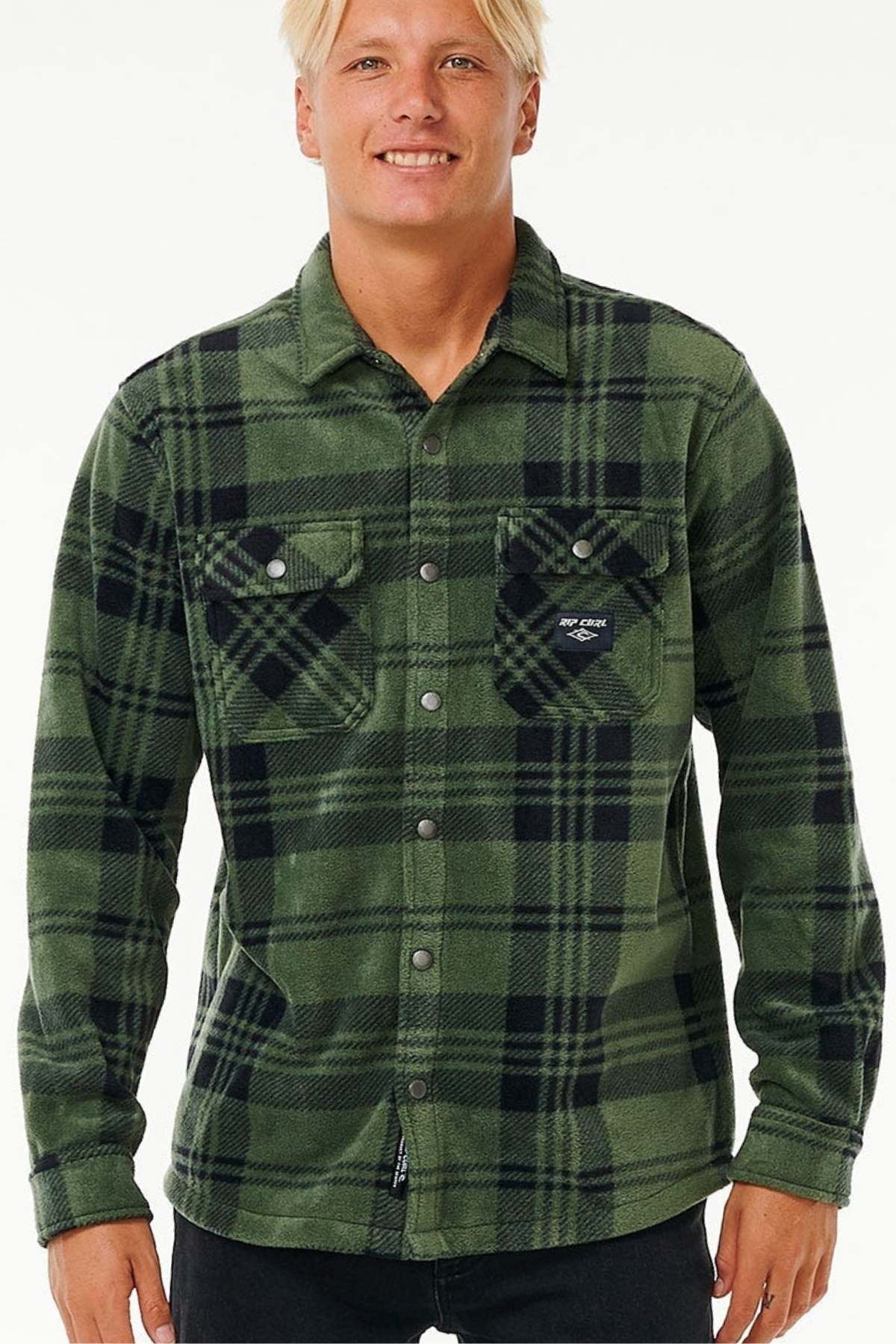 A person with light hair is wearing a green and black plaid Rip Curl Party Pack Polar Fleece Shirt made of recycled polyester, featuring two chest pockets. They have paired it with black pants against a white background.
