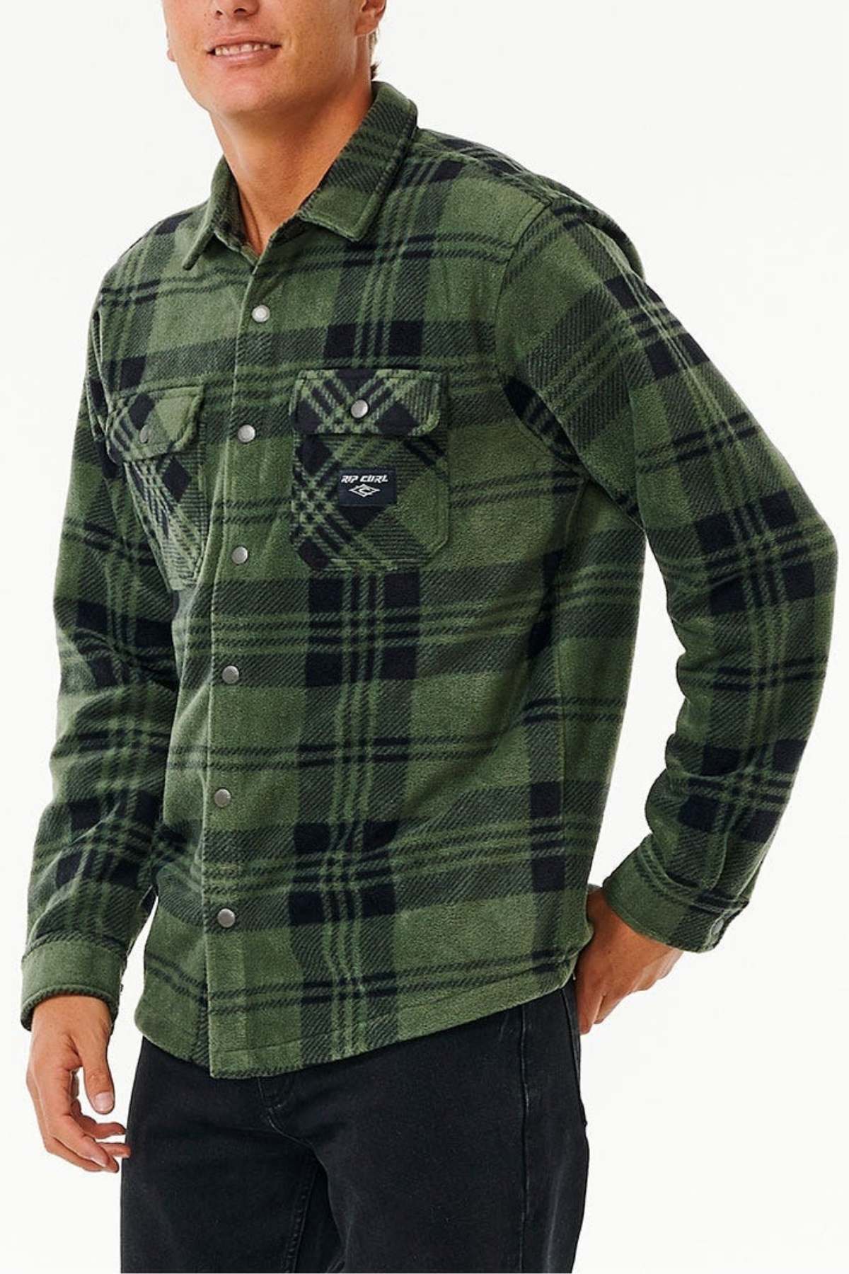 A person wearing a Rip Curl Party Pack Polar Fleece Shirt in green and black plaid, featuring long sleeves, snap buttons, and two chest pockets, is standing with their left hand partially in a pocket of their black pants. The person has short hair and a slight smile against a plain white background.