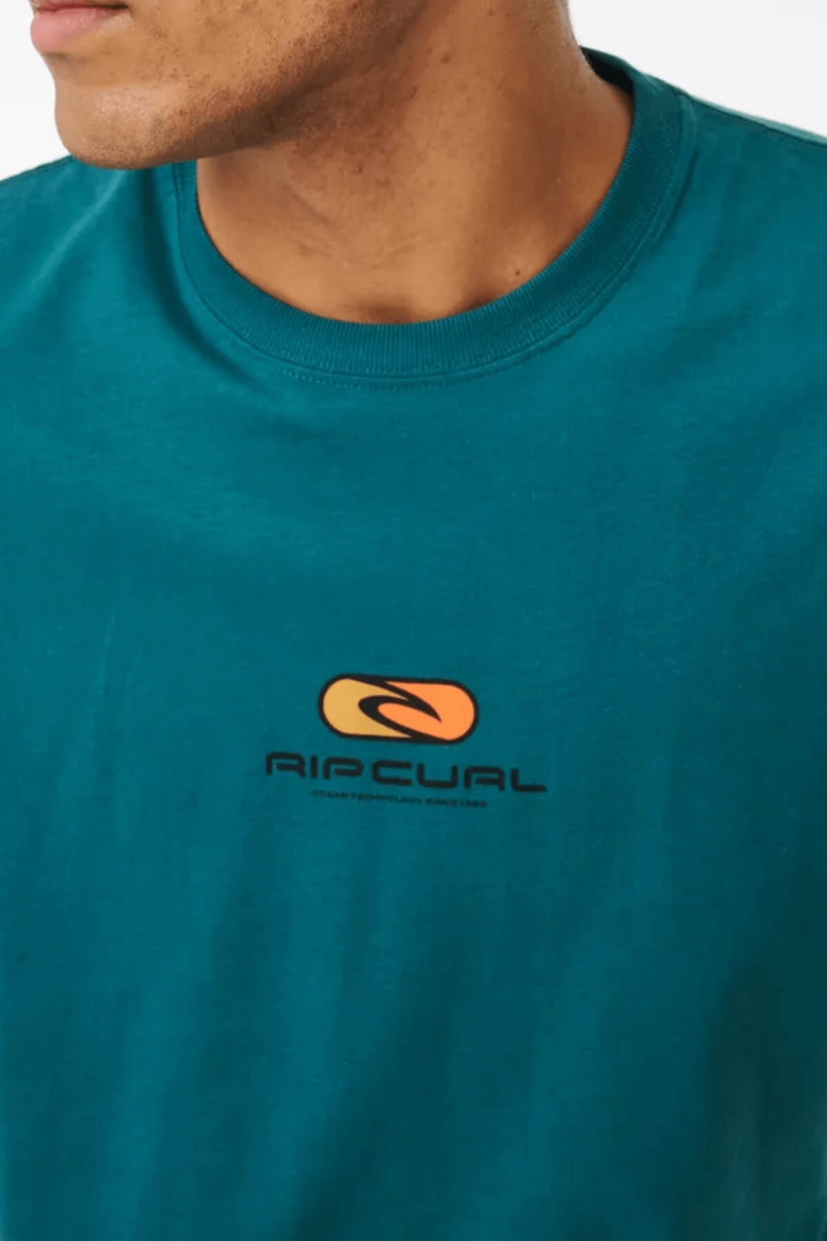 Close-up of a person wearing the Rip Curl Pill Icon Tee, made from 100% cotton in teal. The t-shirt sports a small orange and black logo on the chest with "RIP CURL" text. The focus is on the stylish garment, with the wearer's face partially visible.