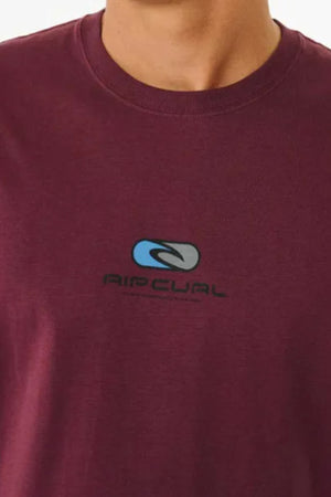 A person in a maroon Rip Curl Pill Icon Tee, featuring a blue and gray logo with "RIP CURL" text on the front. Only their neck and upper chest are visible.