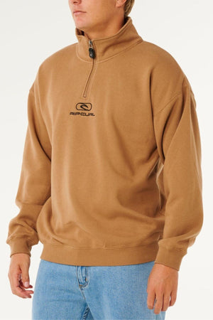 A person wearing a tan Rip Curl Pill Icon 1/4 Zip Crew men's sweatshirt made from organic cotton fleece, paired with light blue jeans, stands against a plain white background.