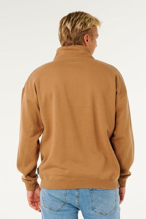 A person with short blond hair is seen from behind, wearing the Rip Curl Pill Icon 1/4 Zip Crew—an organic cotton fleece sweatshirt in light brown—and light blue jeans, set against a plain light background.