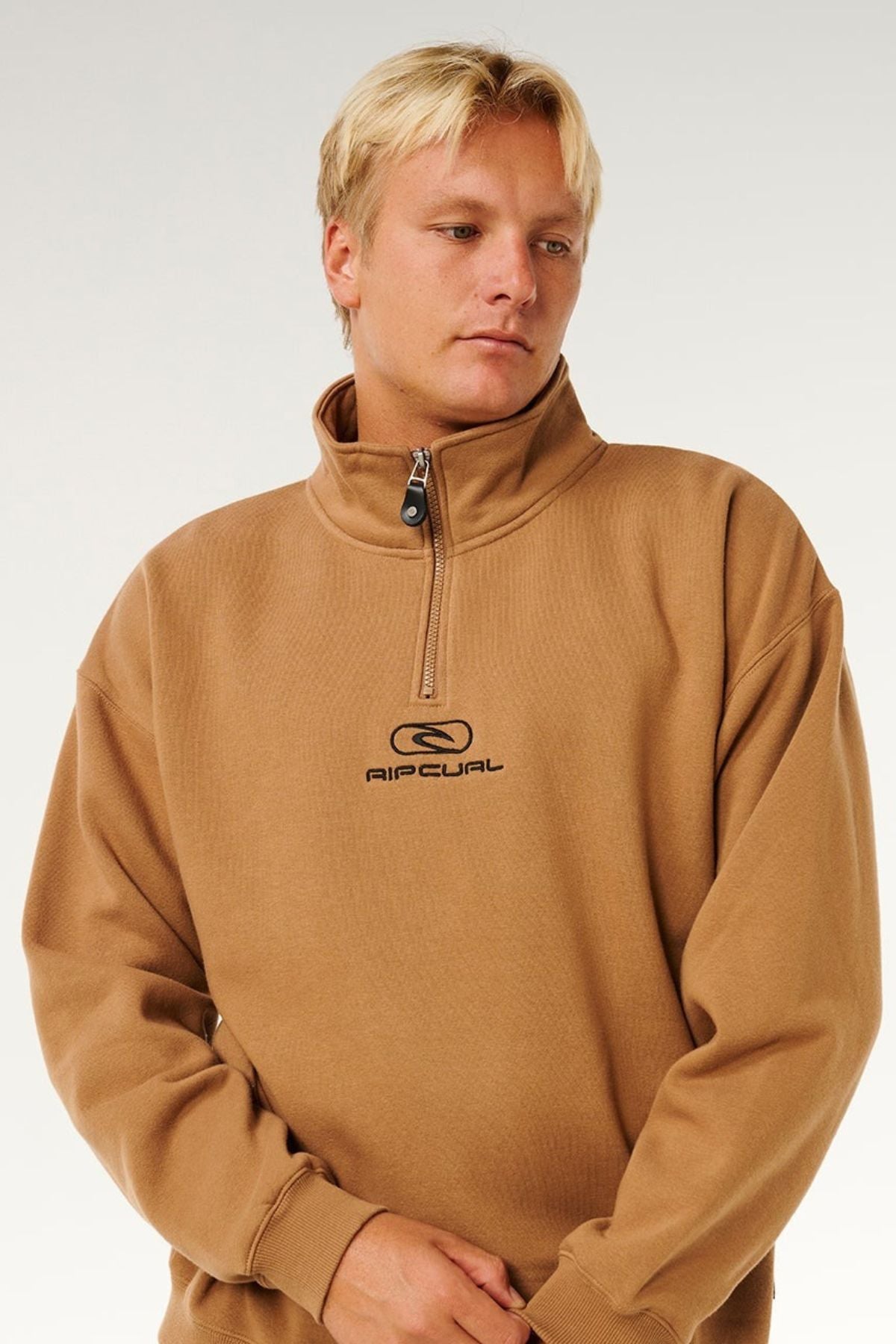A person with short blond hair wears a tan Rip Curl Pill Icon 1/4 Zip Crew men's sweatshirt made from organic cotton fleece, standing against a plain light background and looking slightly to the side with a neutral expression.