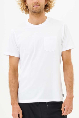 A man with curly hair stands against a white background, wearing a Rip Curl Plain Pocket Tee made from 100% cotton. The T-shirt's standard fit and minimalist design complement his black pants, creating a relaxed yet stylish look.