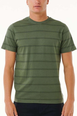 A person wearing a Rip Curl Plain Stripe Tee in green with short sleeves is standing against a plain background. This 100% cotton T-shirt features horizontal black stripes, and the individual pairs it with dark pants.