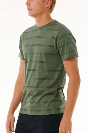 A person dressed in a Rip Curl Plain Stripe Tee, characterized by its green color and short sleeves and crafted from 100% cotton, paired with dark pants, stands against a plain white background, facing slightly to the left.