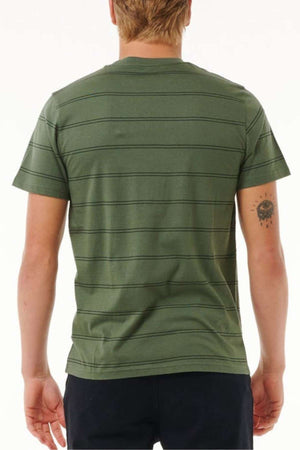 A person with short blond hair is turned away, dressed in a Rip Curl Plain Stripe Tee made entirely from cotton. This green shirt showcases thin black horizontal stripes and is styled with dark shorts. Their right arm displays a tattoo of an eye with lines radiating below, set against a simple white background.