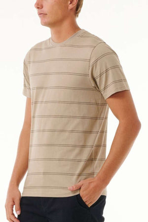 A person wearing a beige, short-sleeved Rip Curl Plain Stripe Tee with black pants stands slightly turned to the left, one hand in their pocket. The background is plain white.