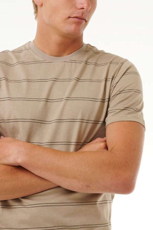 A person stands with arms crossed, wearing the Rip Curl Plain Stripe Tee in beige and black. The background is plain white, and the photo captures the individual from the shoulders down, without showing their face.