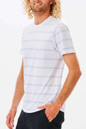 A person with curly hair dons the Rip Curl Plain Stripe Tee, celebrated for its supreme comfort. The T-shirt features a white base adorned with thin blue stripes and pairs seamlessly with black pants. The individual is positioned against a simple white backdrop, wearing a subtle smile and facing slightly to the side.