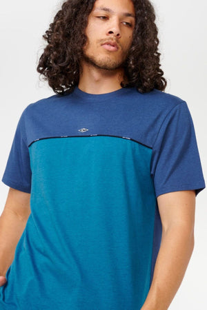 A person with long curly hair is wearing the Rip Curl Vaporcool Medina Seacell Tee in blue and teal. They are standing against a plain background, looking slightly to the side, showcasing the tee’s excellent moisture control.