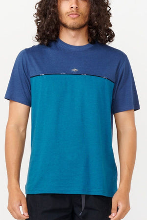 A person with long curly hair is wearing a Rip Curl Vaporcool Medina Seacell Tee in two shades of blue and black pants, standing against a plain background. The T-shirt has a dark blue top and a lighter blue bottom, adorned with a small logo in the center that provides moisture control.