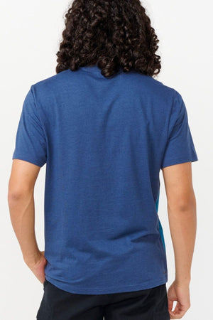 A person with long curly hair stands with their back to the camera, wearing a Rip Curl Vaporcool Medina Seacell Tee and black pants. The light-colored background highlights the stylish tee, known for its moisture control features.