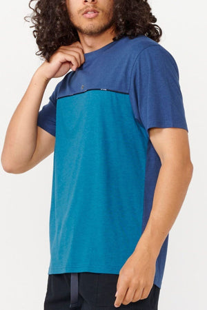 A person with curly hair is wearing a Rip Curl Vaporcool Medina Seacell Tee, showcasing blue and teal color-blocking, paired with black pants. They are facing away from the camera against a light backdrop, enjoying the tee's moisture control feature.