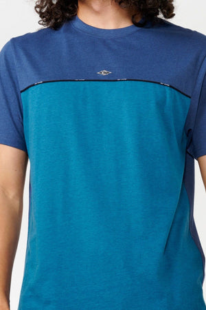 A person with curly hair is wearing a Rip Curl Vaporcool Medina Seacell Tee in two-tone blue, showcasing a small logo on the chest. The shirt has a dark blue upper section and a lighter blue lower section, divided by a thin horizontal line. The background is plain white.