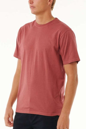 A person wearing a Rip Curl Plain Wash Tee in a casual maroon shade and dark pants, epitomizing laid-back fashion, stands against a neutral background.