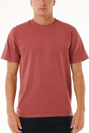 A person is dressed in a Rip Curl Plain Wash Tee, a minimalist maroon cotton top, paired with black pants against a light background, perfectly capturing the essence of casual fashion. The emphasis is on the upper part of the outfit.