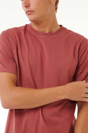 A person wearing the Rip Curl Plain Wash Tee, a short-sleeved maroon cotton shirt, stands against a neutral background with one arm crossed over the other. The person's head is turned slightly to the side, embodying effortless casual fashion.