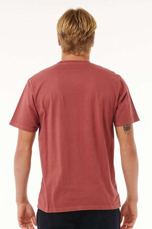 A person with blond hair is facing away, dressed in a short-sleeved, rusty red Rip Curl Plain Wash Tee made of cotton and dark pants. The background is plain white, and part of a small tattoo is visible on their left arm, exuding an effortlessly casual fashion vibe.