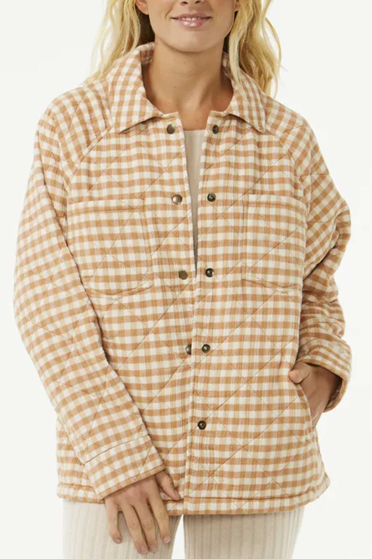 A woman with long blonde hair stands against a plain light background, wearing the Rip Curl Premium Quilted Check Jacket in beige and white, featuring snap buttons, a collar, side pockets, and soft Cotton Double Weave Gauze.
